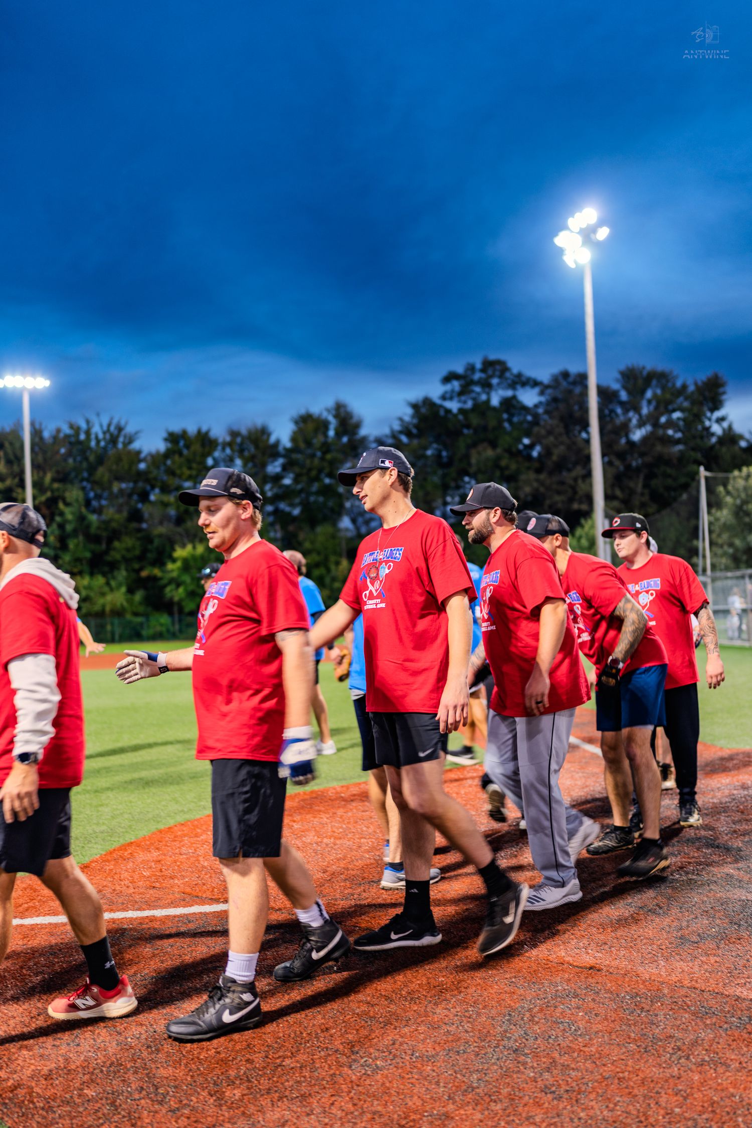 Group of men in red t-shirts and caps walking on a baseball field under bright stadium lights at dusk.