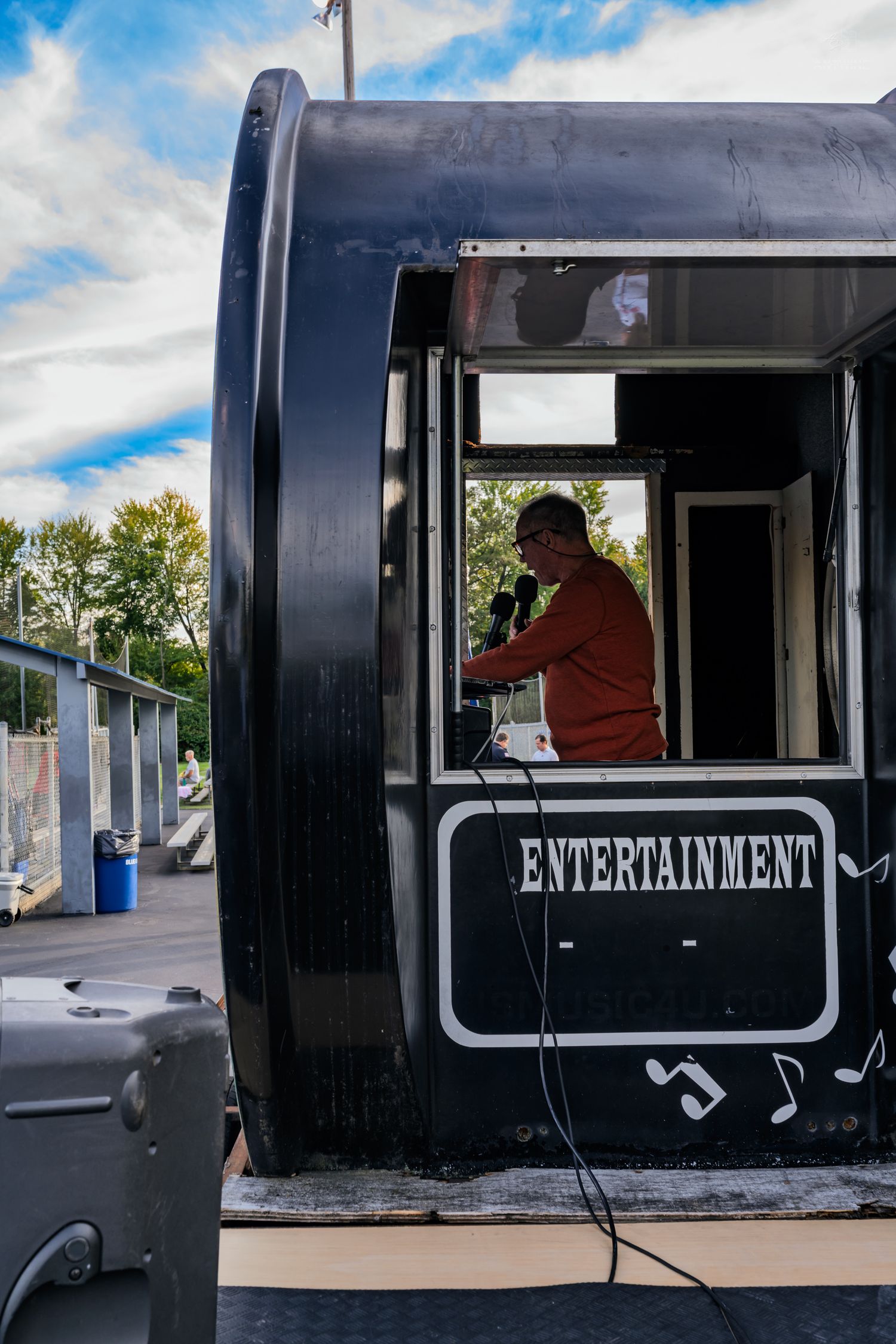 A person speaks into a microphone inside a black entertainment booth outdoors under a partly cloudy sky.