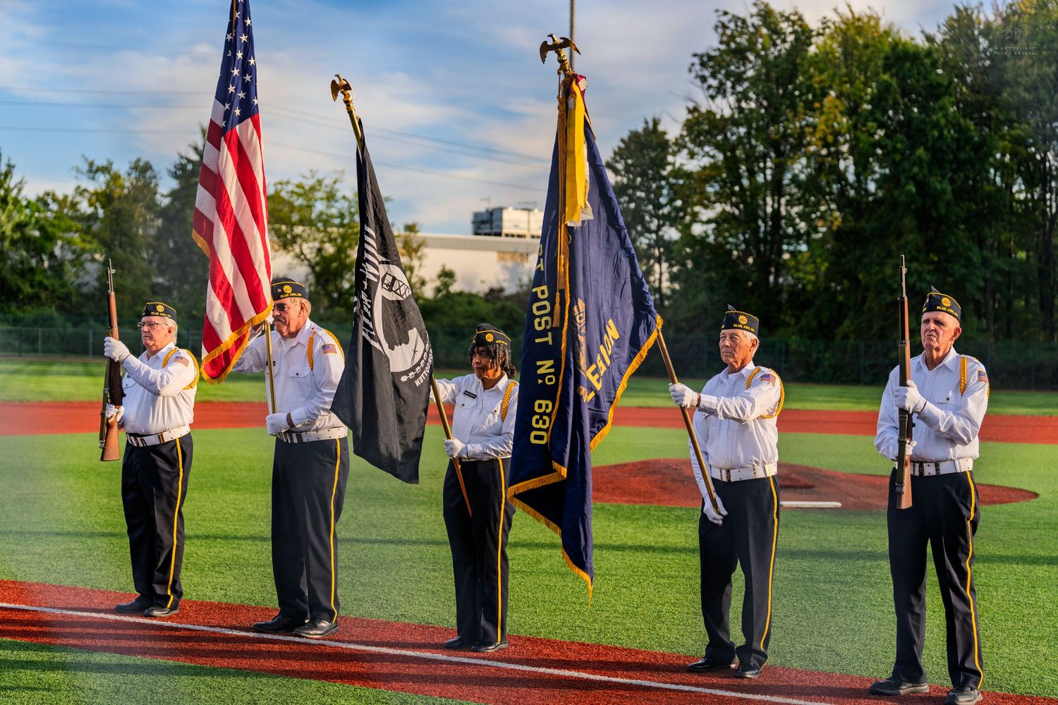 Military color guard presents American and POW flags on a baseball field at an evening ceremony.
