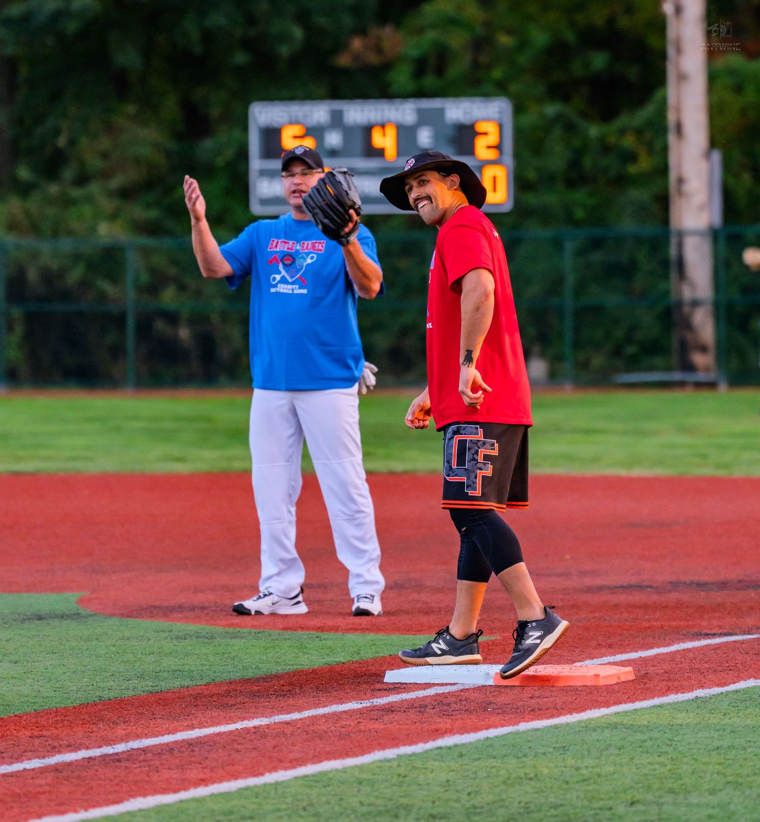 Two softball players near base, one in blue shirt gesturing while other in red stands on base.
