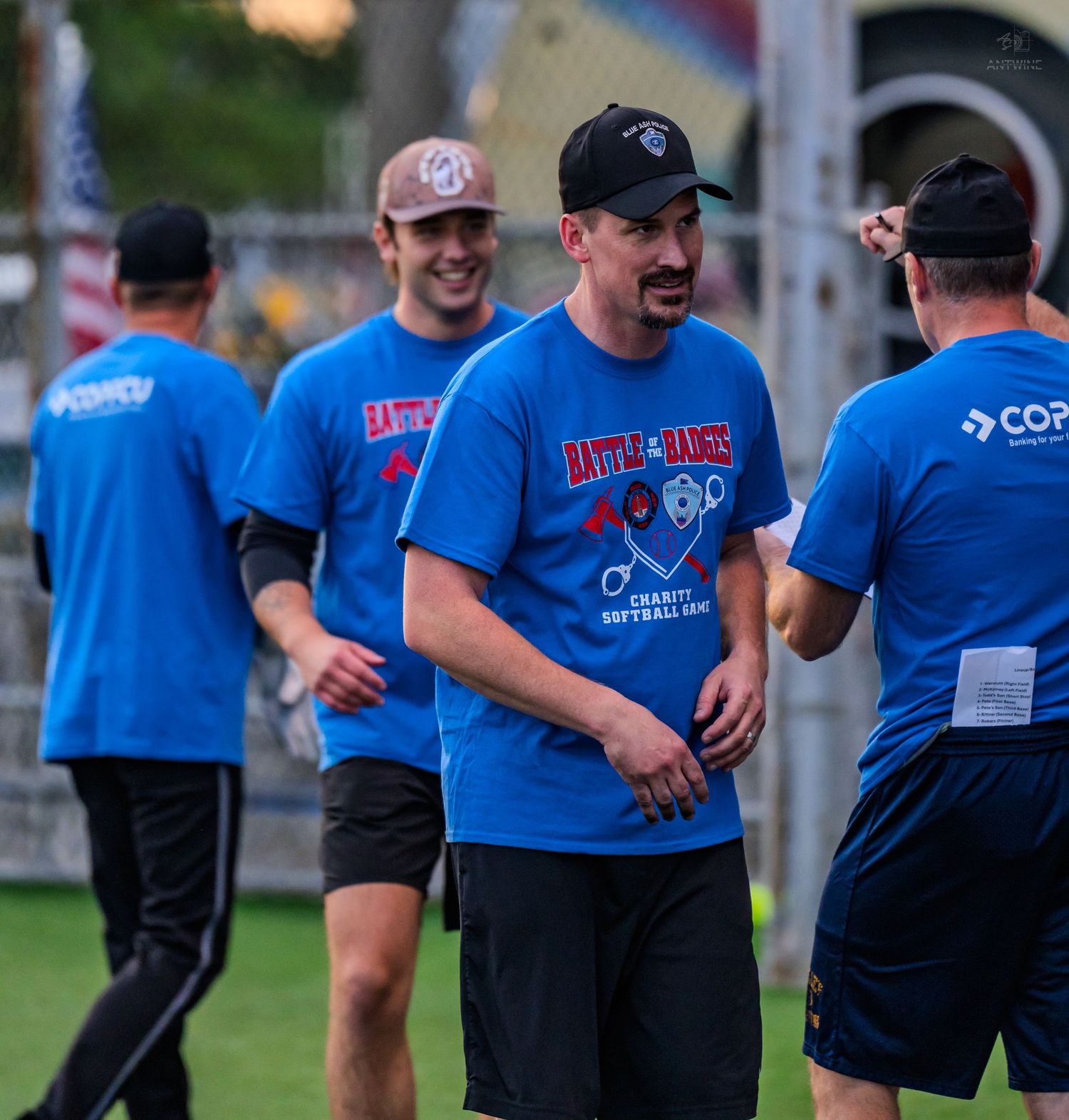 Men in blue Battle of the Badges charity softball game t-shirts socializing on outdoor sports field.
