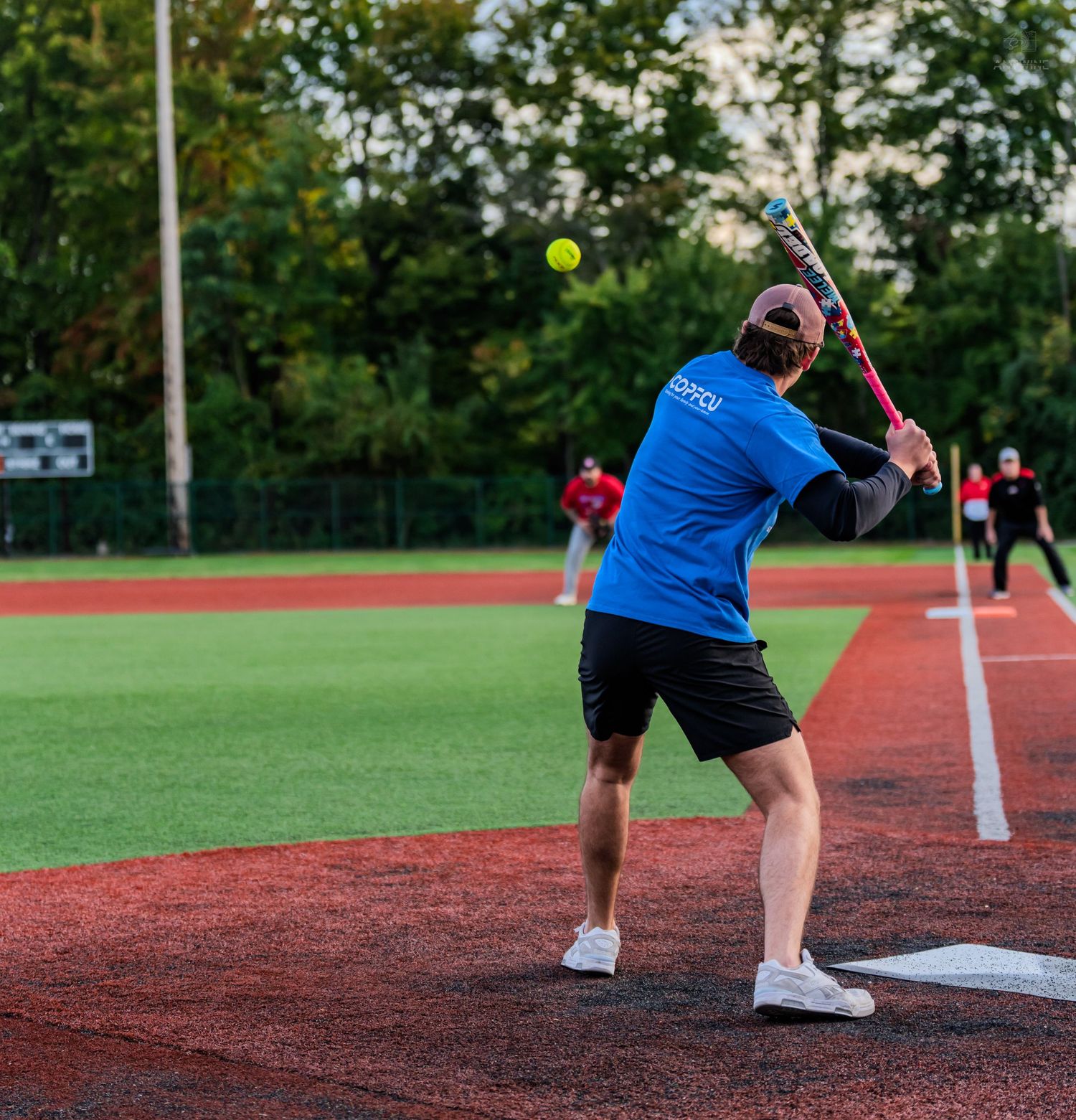 Softball player in blue shirt swings bat at yellow ball during an outdoor game on red turf field.