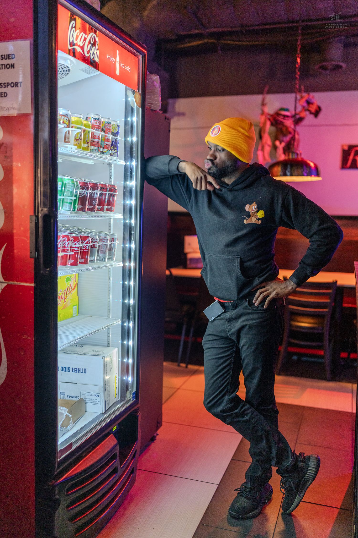 Man in yellow beanie leans against illuminated Coca-Cola beverage cooler in neon-lit restaurant.