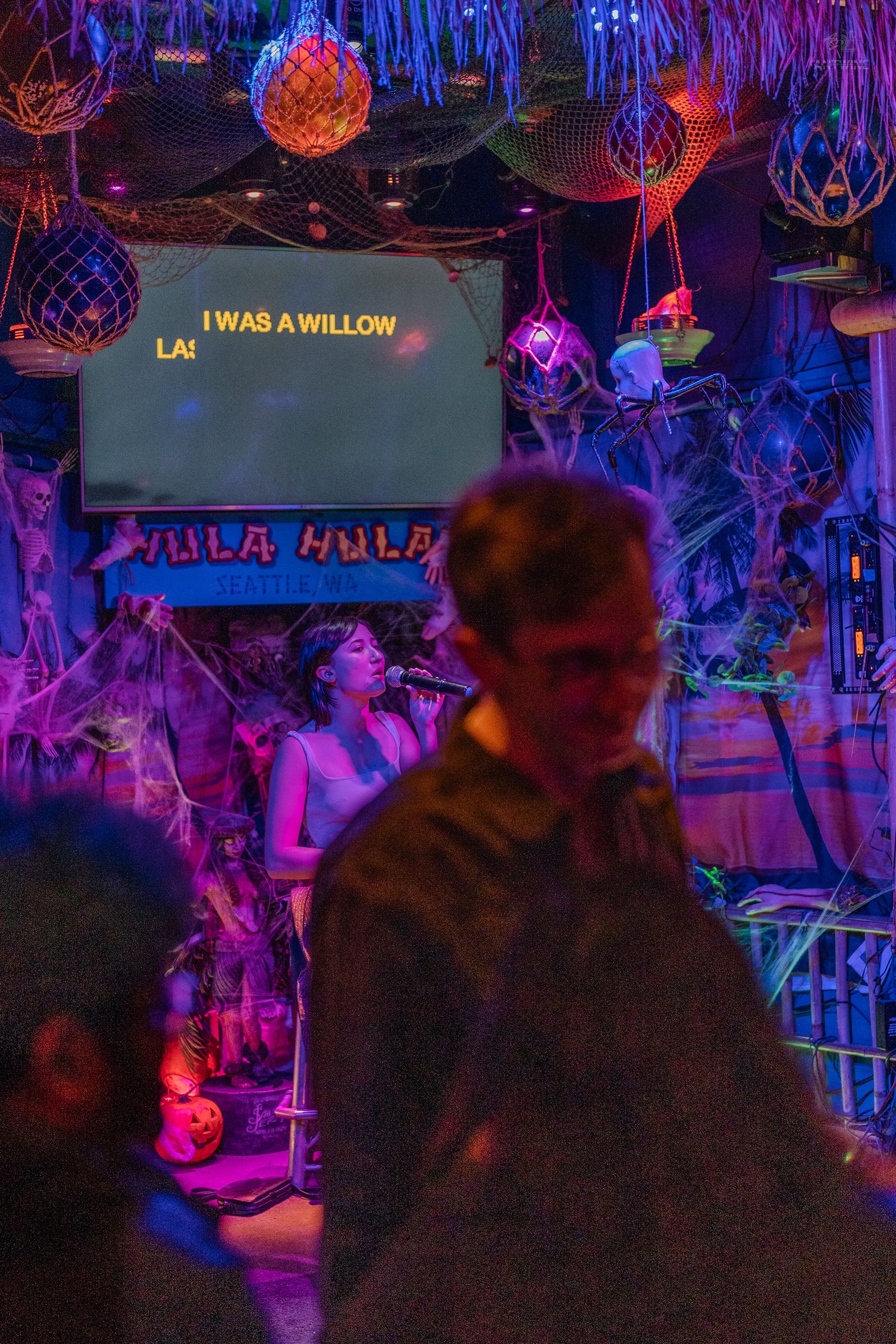 Woman singing karaoke at Hula Hula bar in Seattle with colorful tiki decorations and Halloween cobwebs.