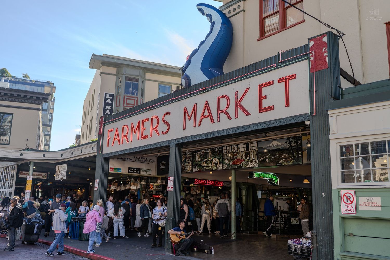 Crowds gather outside the iconic Pike Place Farmers Market in Seattle with a large blue fish sign overhead.