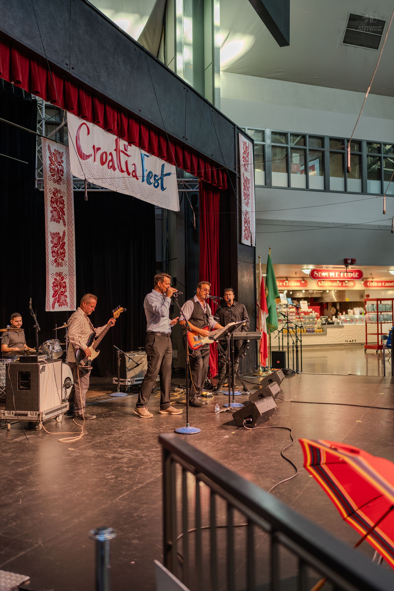 Rock band performs live at CroatiaFest with guitars and microphones on an indoor stage with red curtain backdrop.