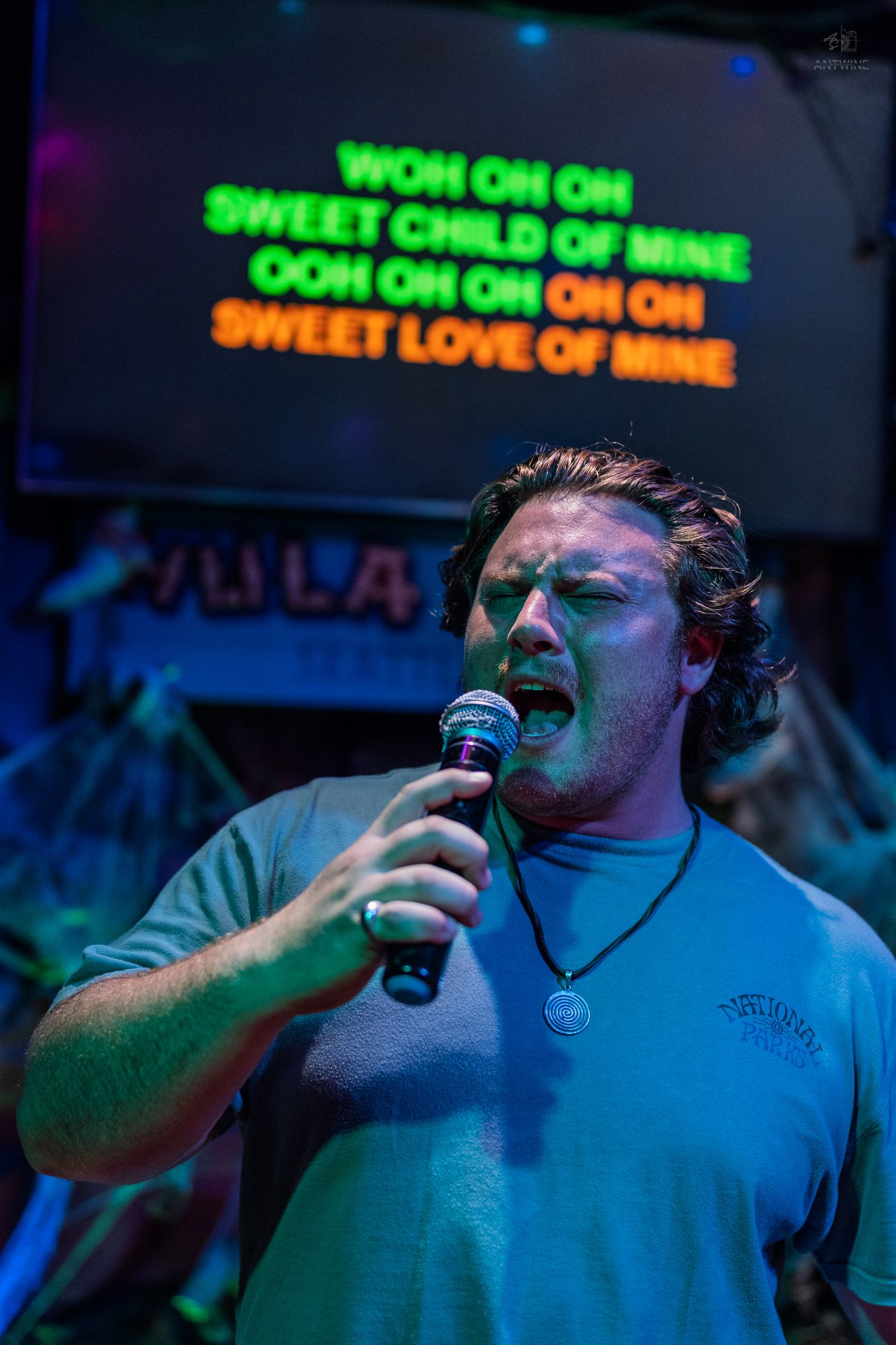 Man singing karaoke to Sweet Child of Mine by Guns N Roses, holding microphone with lyrics displayed on screen behind him.