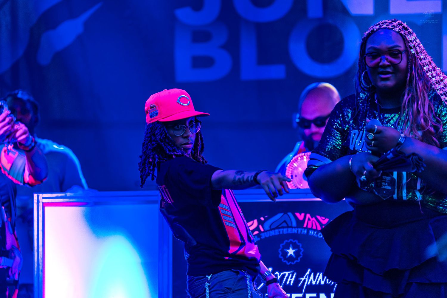 Two performers on stage at Juneteenth Block Party, one in pink cap, under vibrant blue stage lighting.
