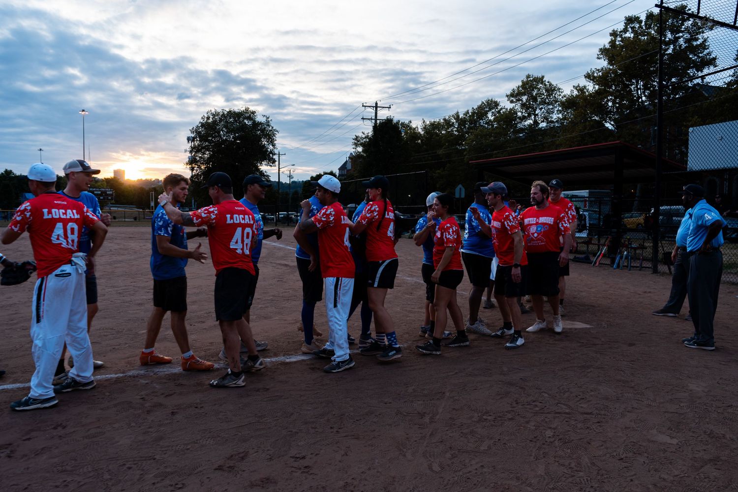 Softball players in red Local jerseys shake hands at sunset after a community league game.