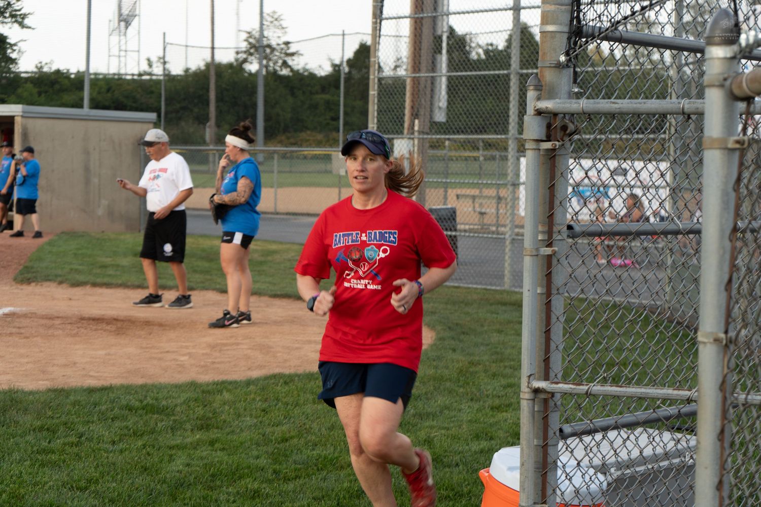 A woman in a red shirt jogging past a chain-link fence at a baseball field during evening practice.