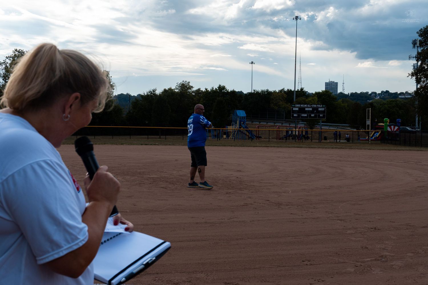 A woman holds a microphone and clipboard at a softball field while a player stands on the dirt infield at dusk.