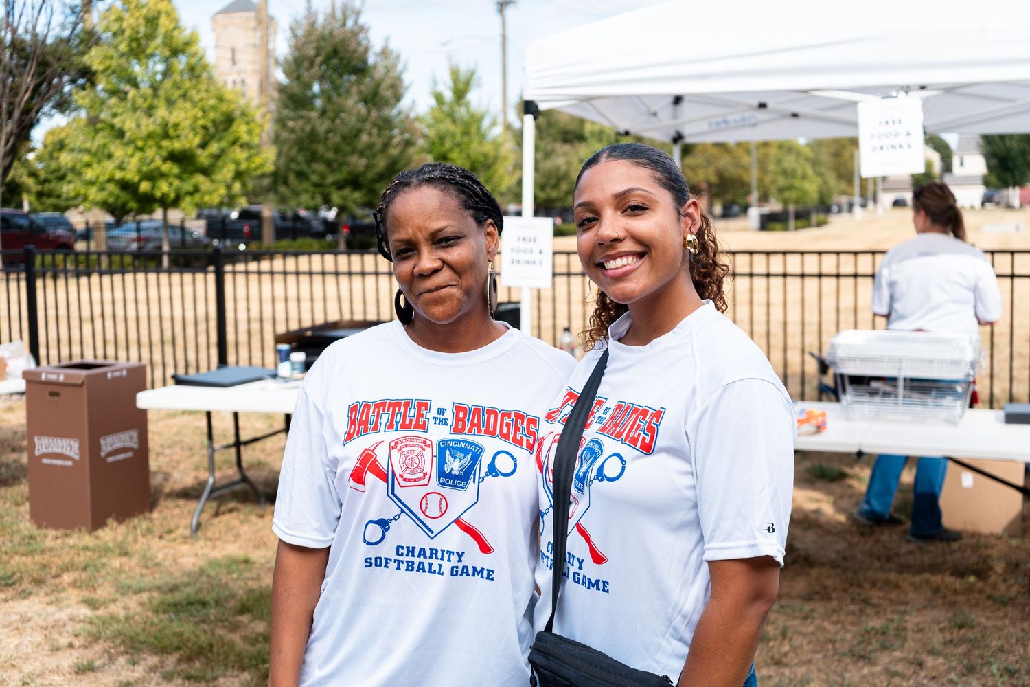 Two women smile wearing charity softball game t-shirts at an outdoor fundraising event on a sunny day.
