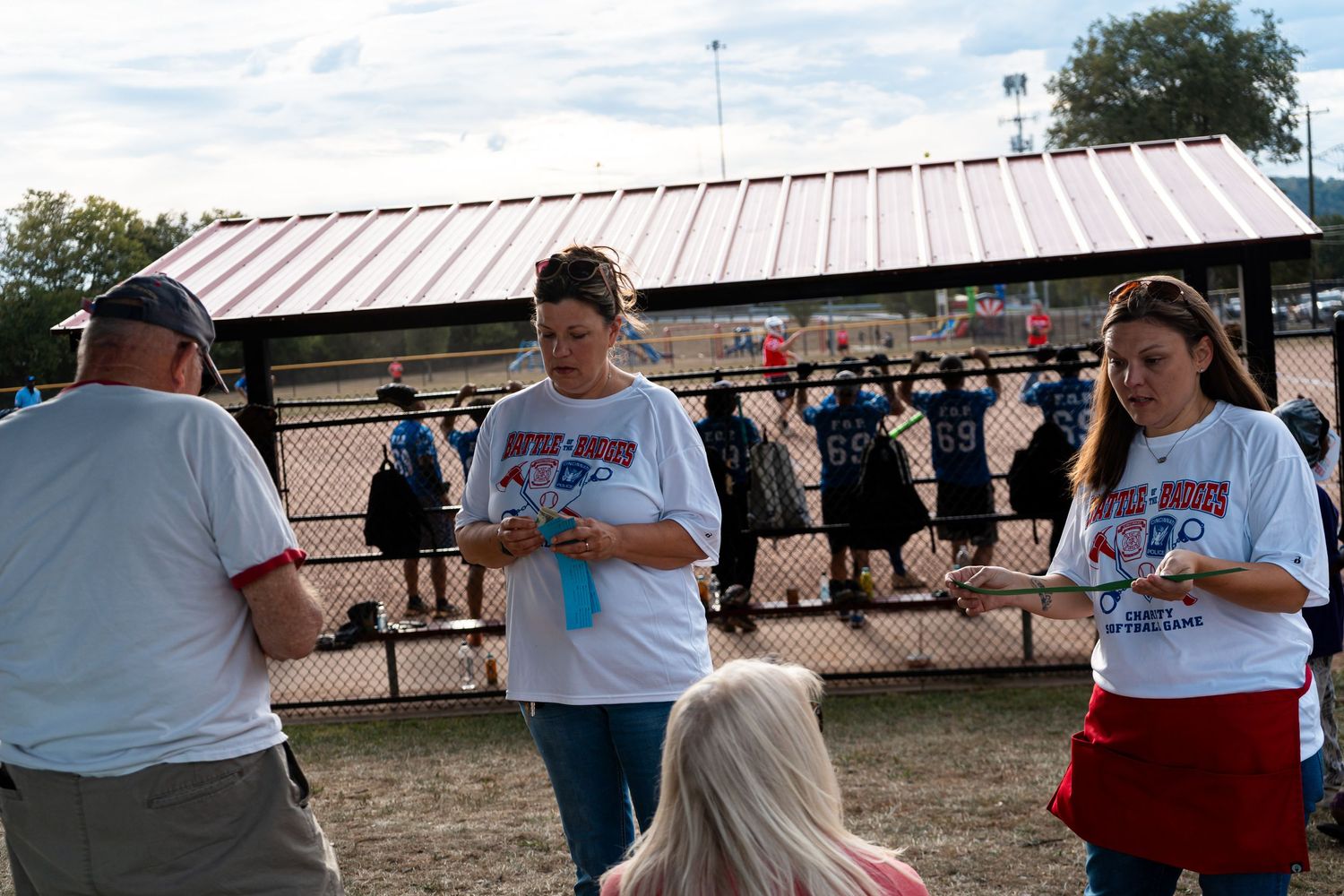 People in matching white t-shirts reviewing papers at an outdoor community event near a fenced sports field.