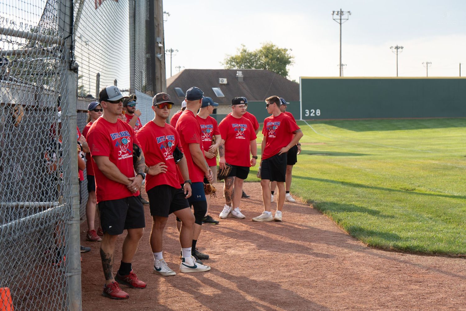 Baseball team in red uniforms standing along dugout fence on a sunny day at a baseball field.