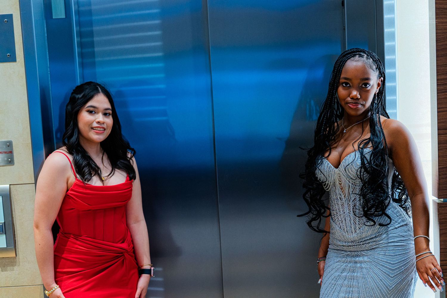 Two young women in formal dresses posing together in front of elevator doors.