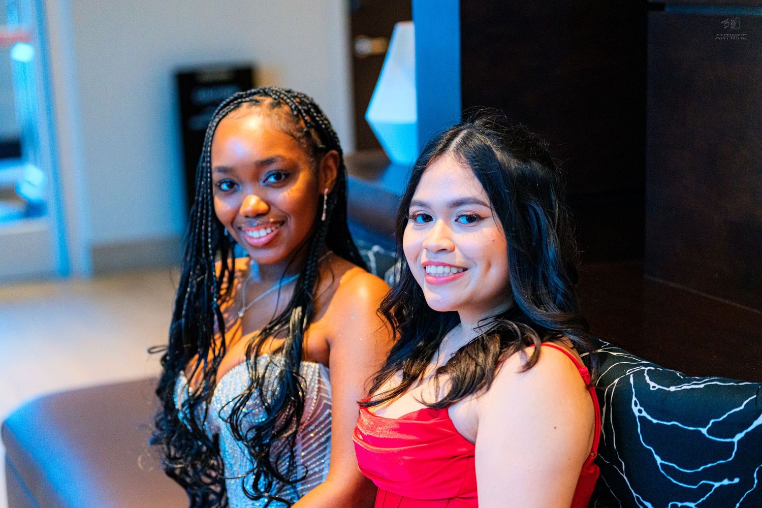 Two smiling young women in formal attire posing closely together at an indoor event.