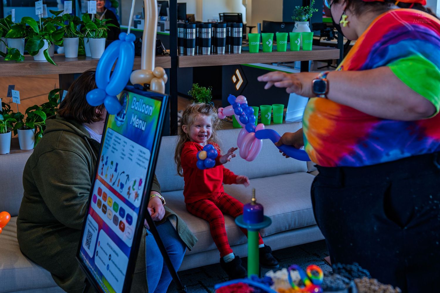 A child in red laughs as a clown in tie-dye creates balloon animals at a colorful indoor event.