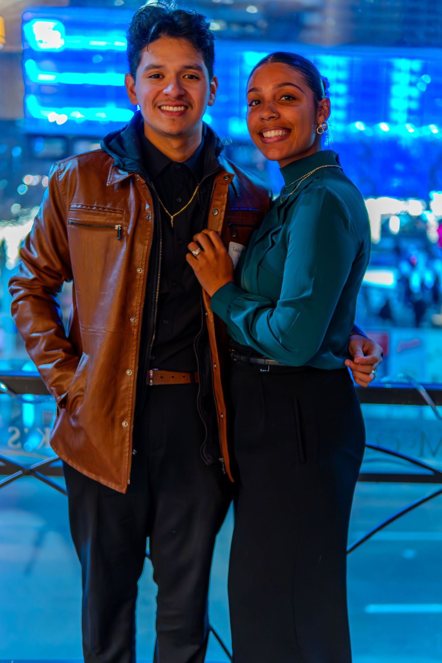 A smiling couple poses together, man in brown leather jacket and woman in teal blouse, against blue neon background.
