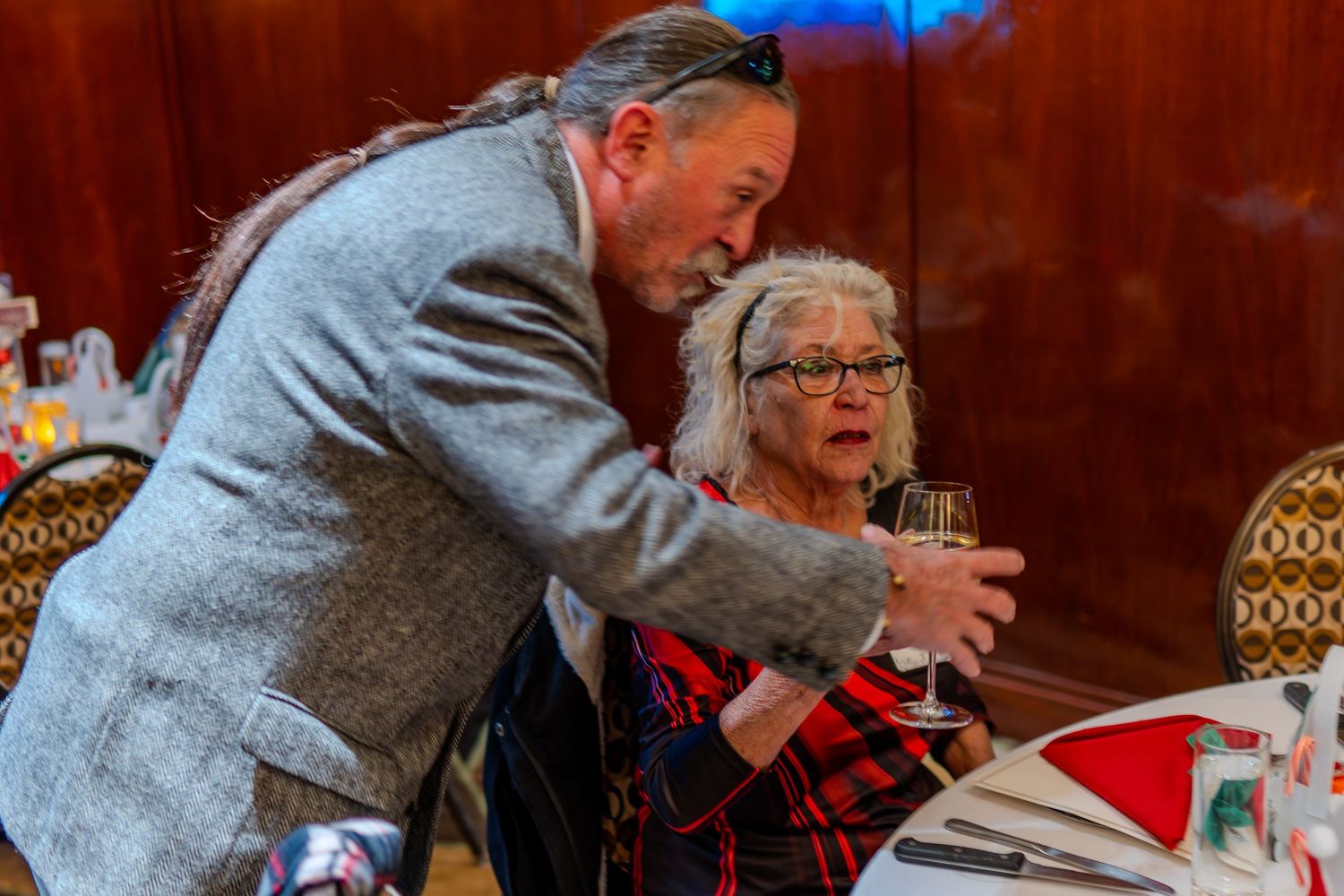 Man in gray blazer leans over elderly woman in red plaid holding champagne at formal dinner event.