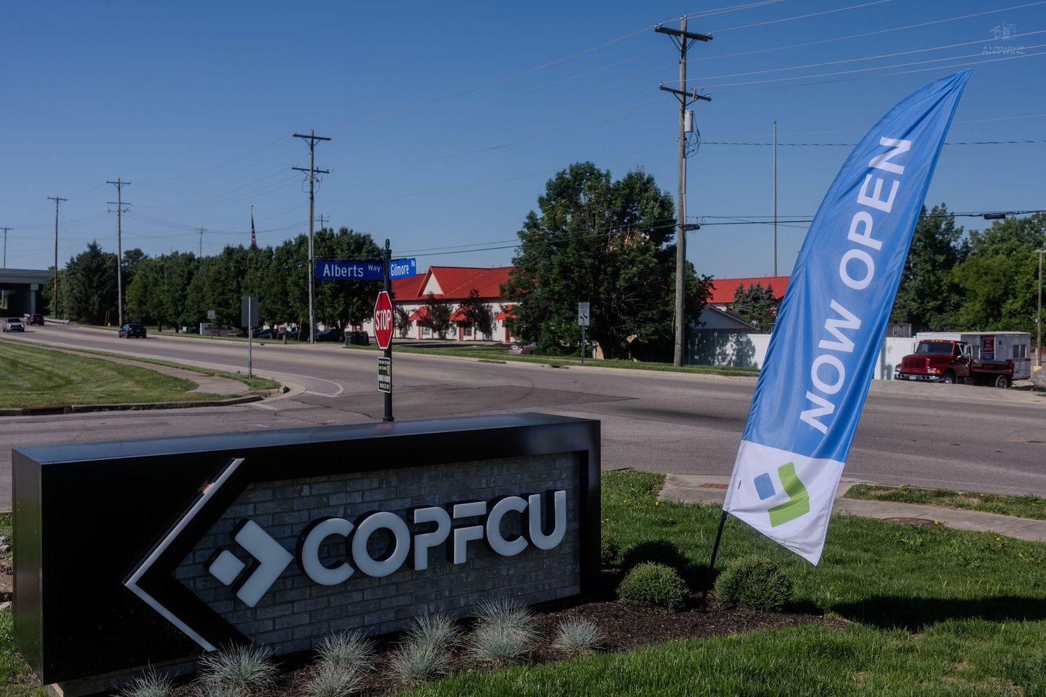 COPFCU credit union sign with a 'Now Open' banner flag near a busy intersection on a sunny day.