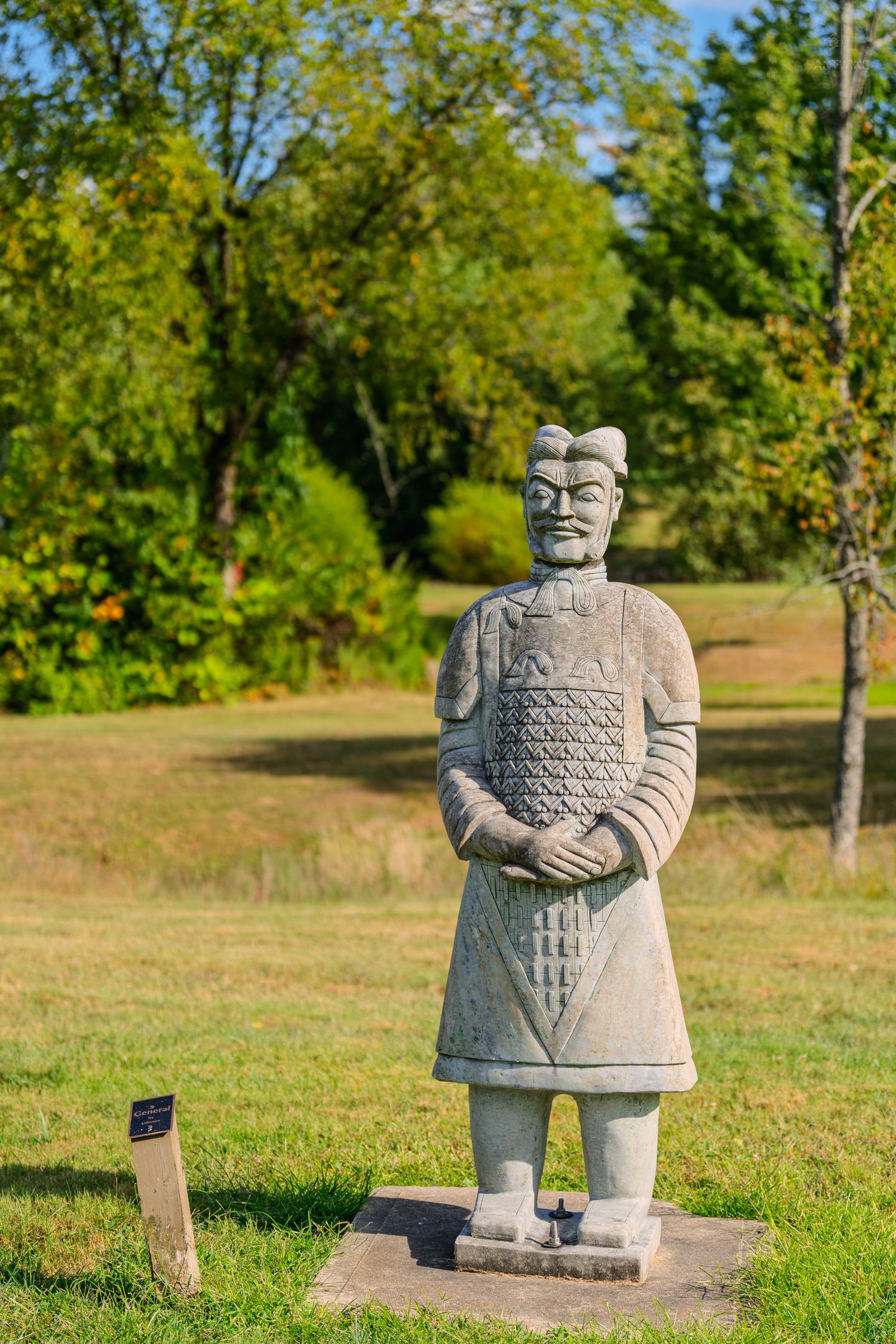 Ancient Chinese terracotta warrior statue replica standing on grass in a lush green park on a sunny day.