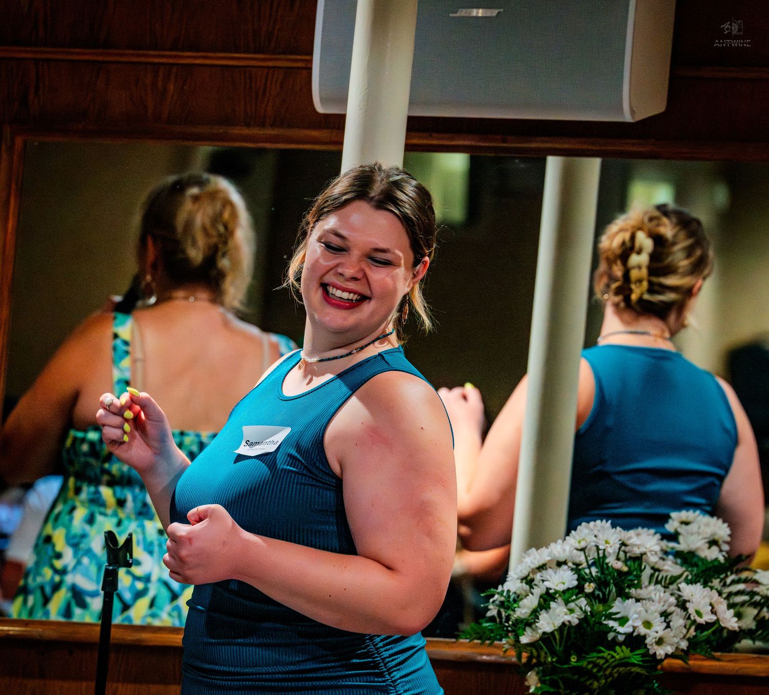 Smiling woman in teal dress poses near mirror at a formal indoor gathering event.