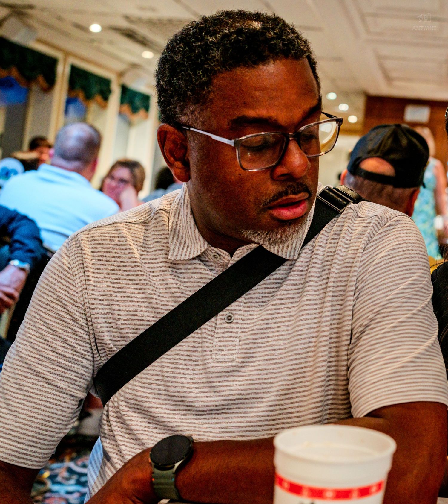 A man wearing glasses and a striped polo shirt with a crossbody bag sits at a busy indoor event.