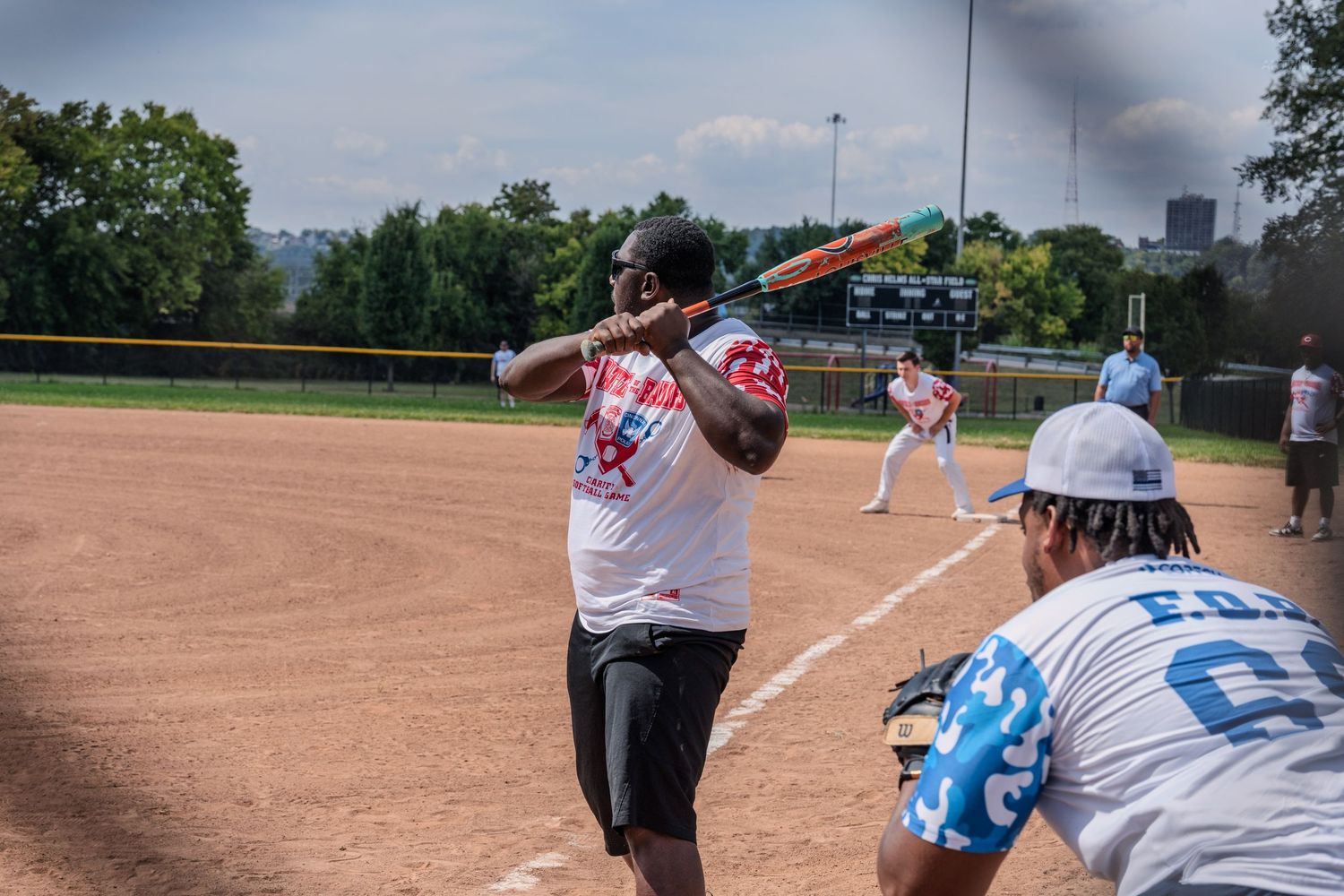 Softball batter in white jersey prepares to swing while a teammate crouches nearby on a sunny day.