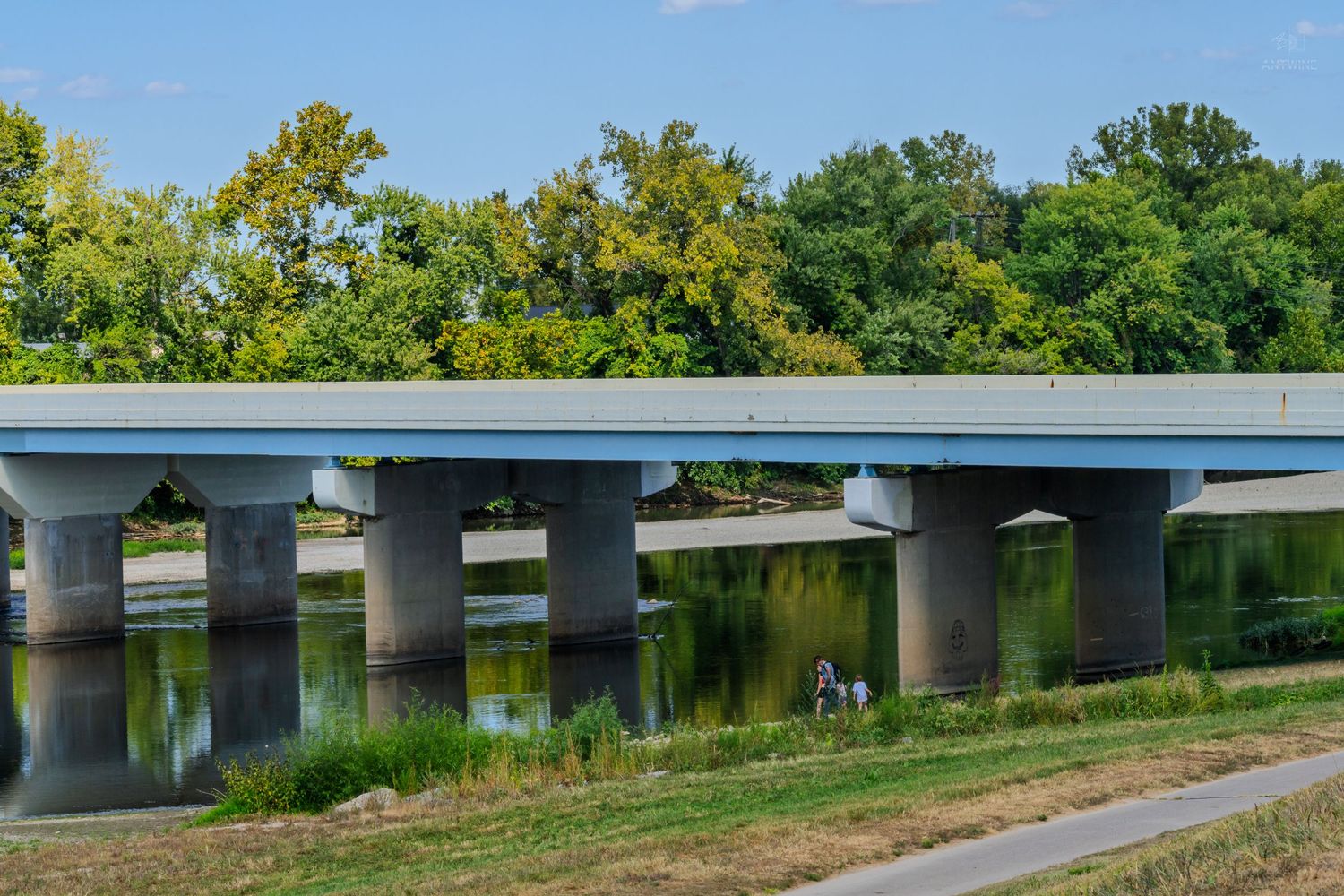 Modern concrete highway bridge spanning a calm river, surrounded by lush green trees on a sunny day.