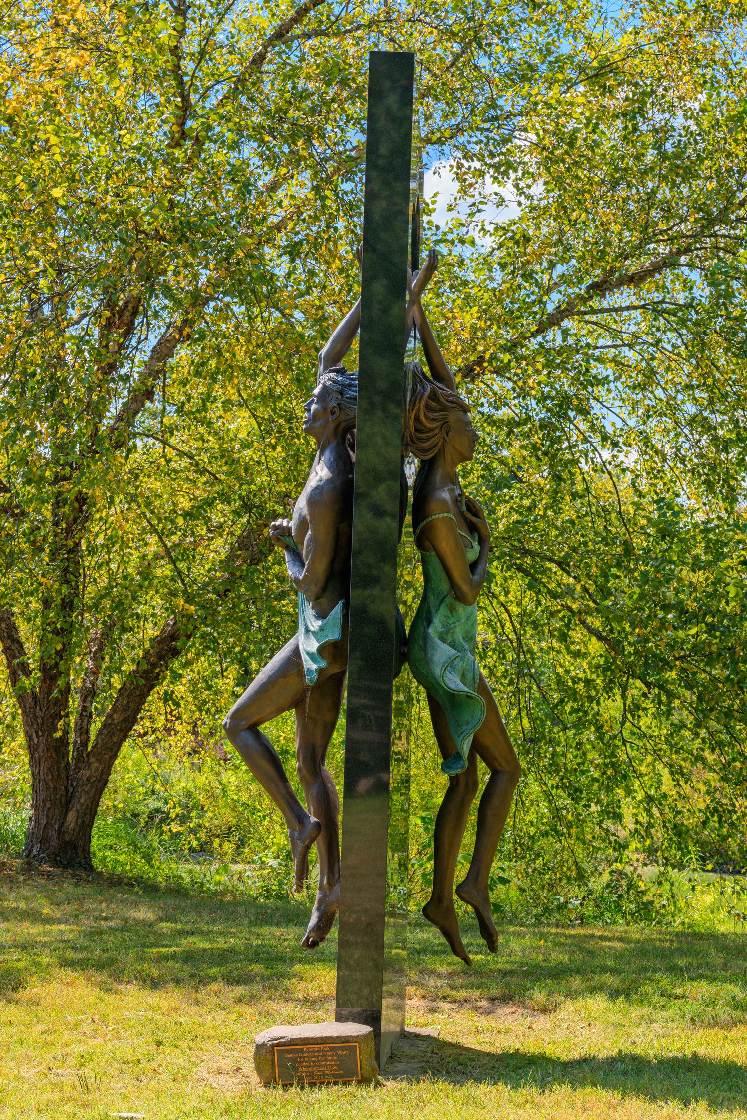Two bronze figures climbing a tall metal column sculpture outdoors surrounded by lush green trees.