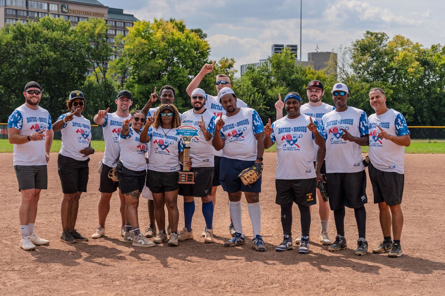 Softball team poses with trophy on dirt field, wearing matching white jerseys on a sunny day.