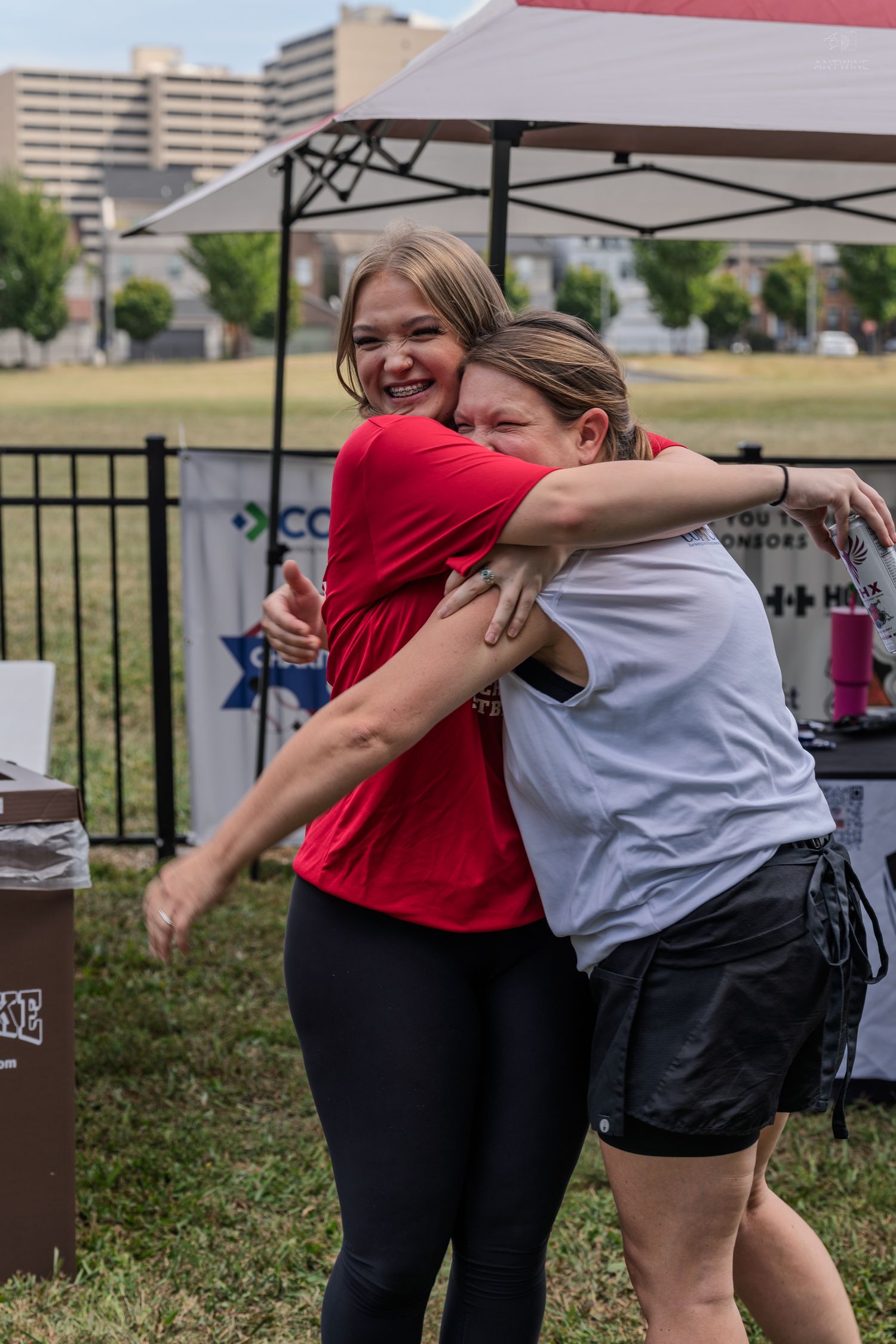 Two women share a joyful hug outdoors at a charity softball game event on a sunny day.