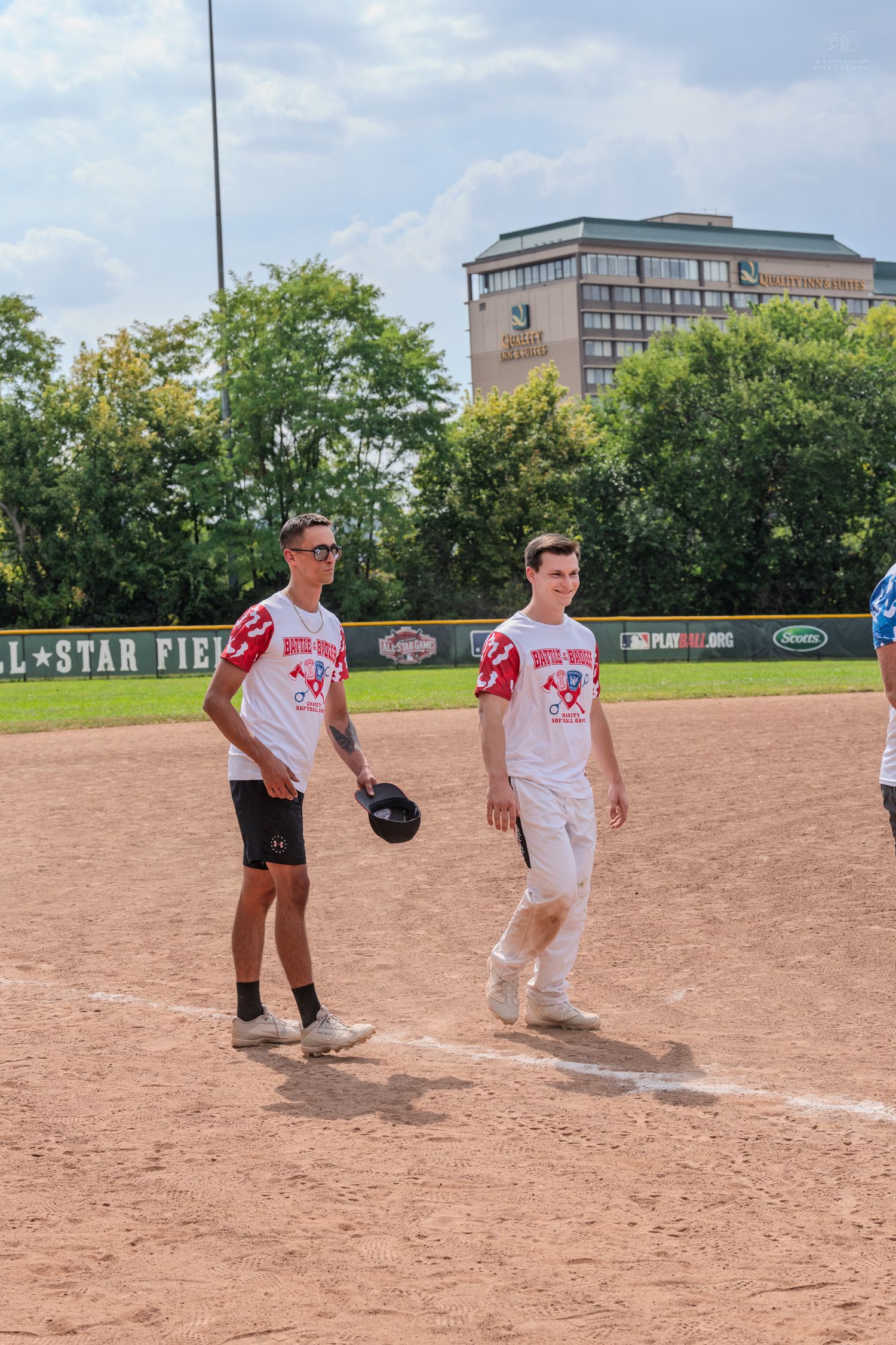 Two young men in matching white softball uniforms walking on a baseball field near first base on a sunny day.