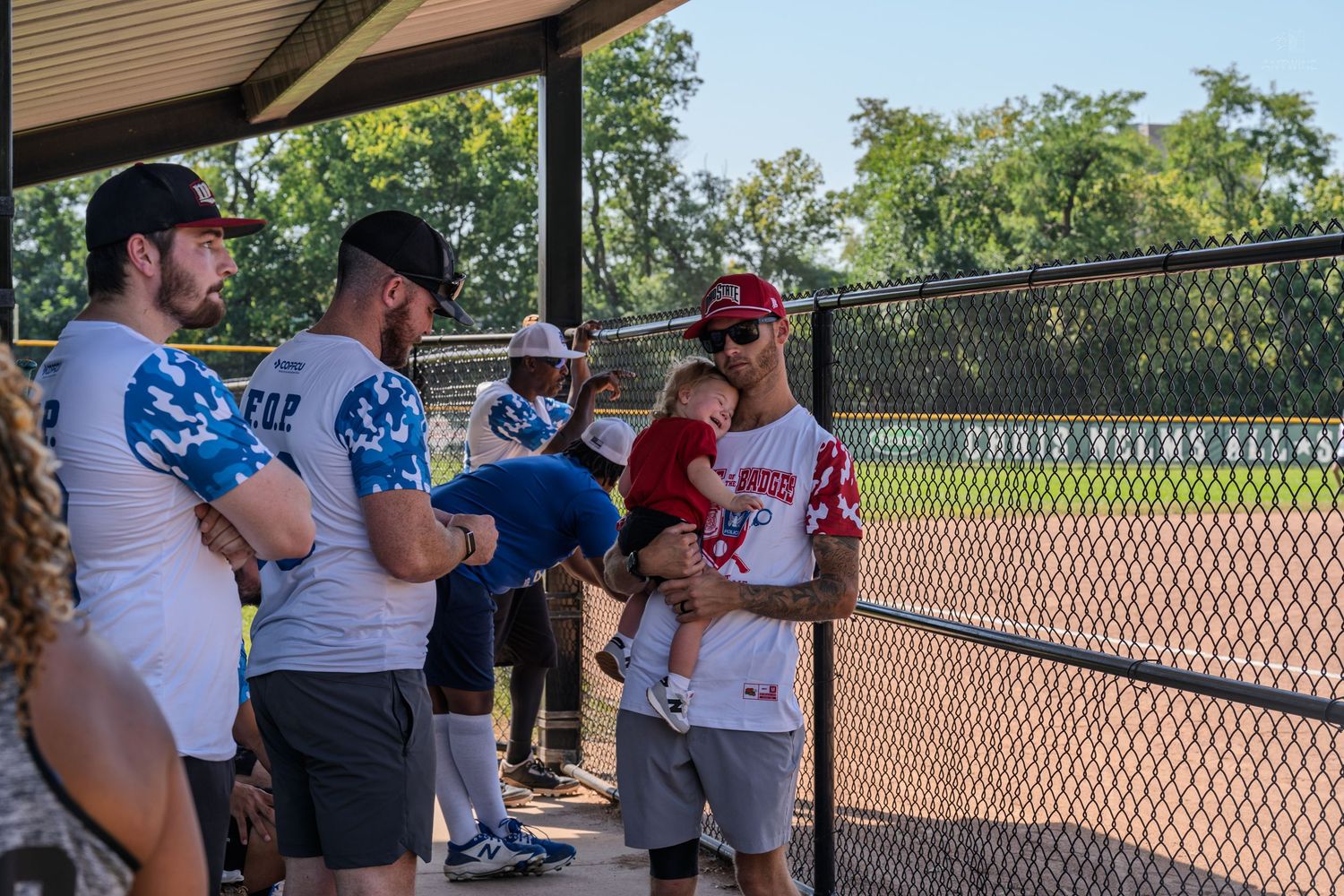 Group of men in baseball uniforms talking in dugout during a sunny outdoor softball game.