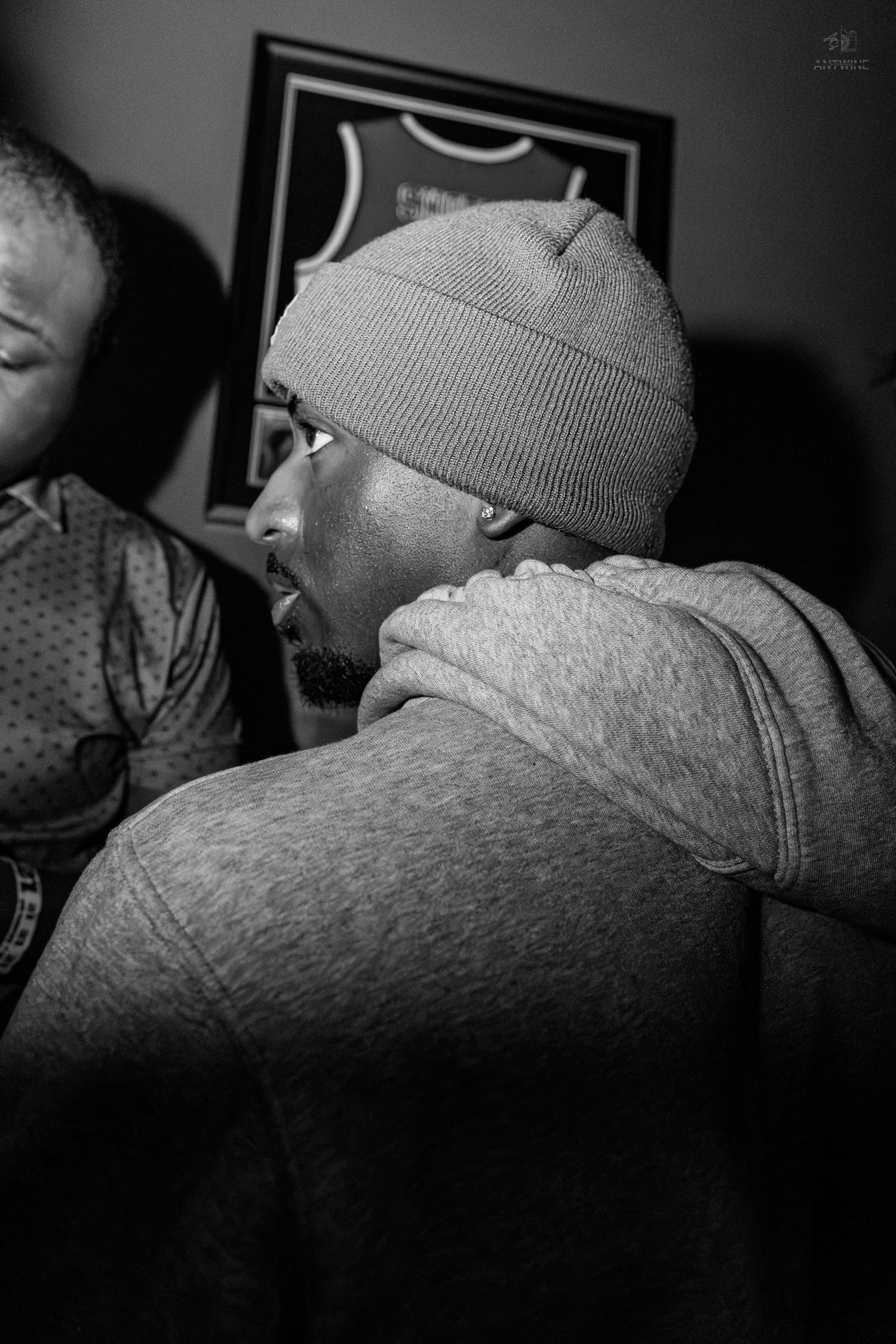Man in grey beanie and hoodie at an indoor gathering, candid black and white portrait.