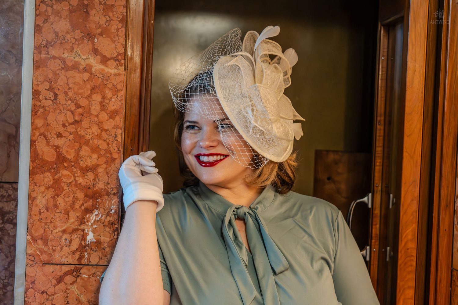 Woman in vintage 1940s style mint dress and cream fascinator hat with veil, smiling with red lips.