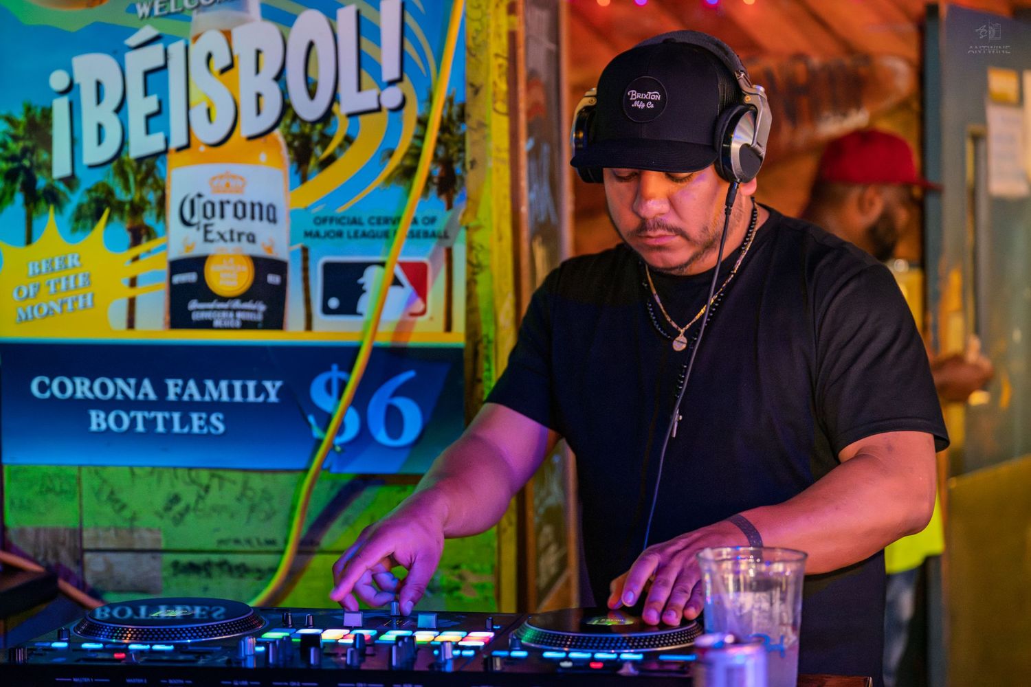DJ mixing tracks at a Corona Extra-sponsored baseball event, wearing headphones and a black cap.