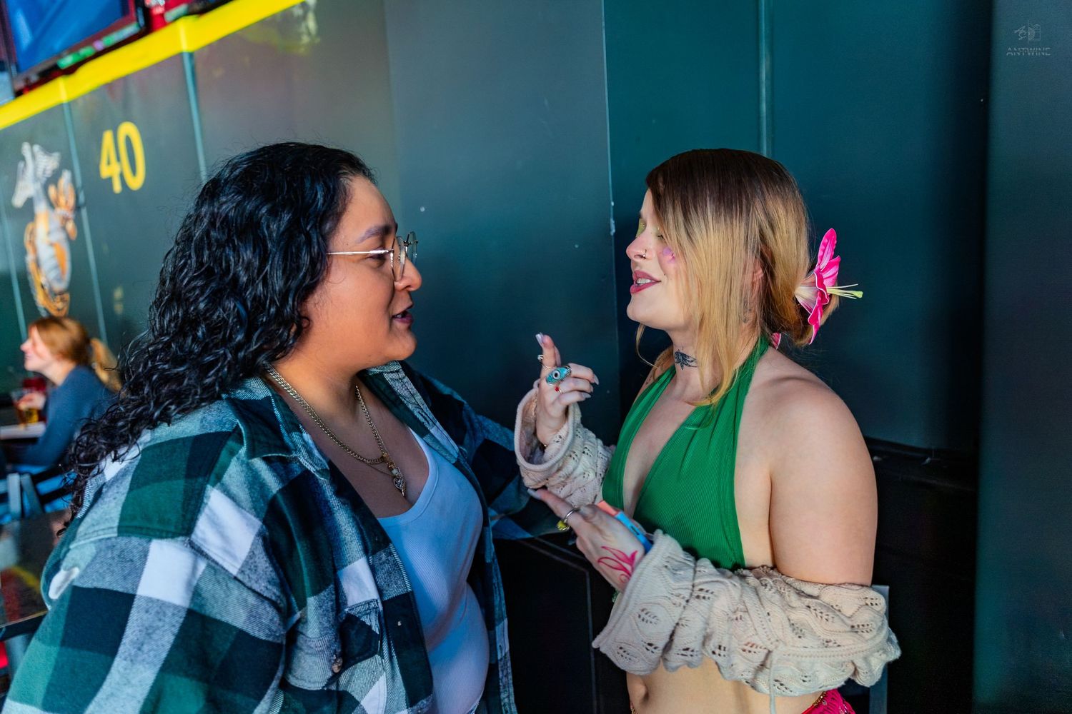 Two women in festive costumes chat animatedly at an indoor event near a green wall.