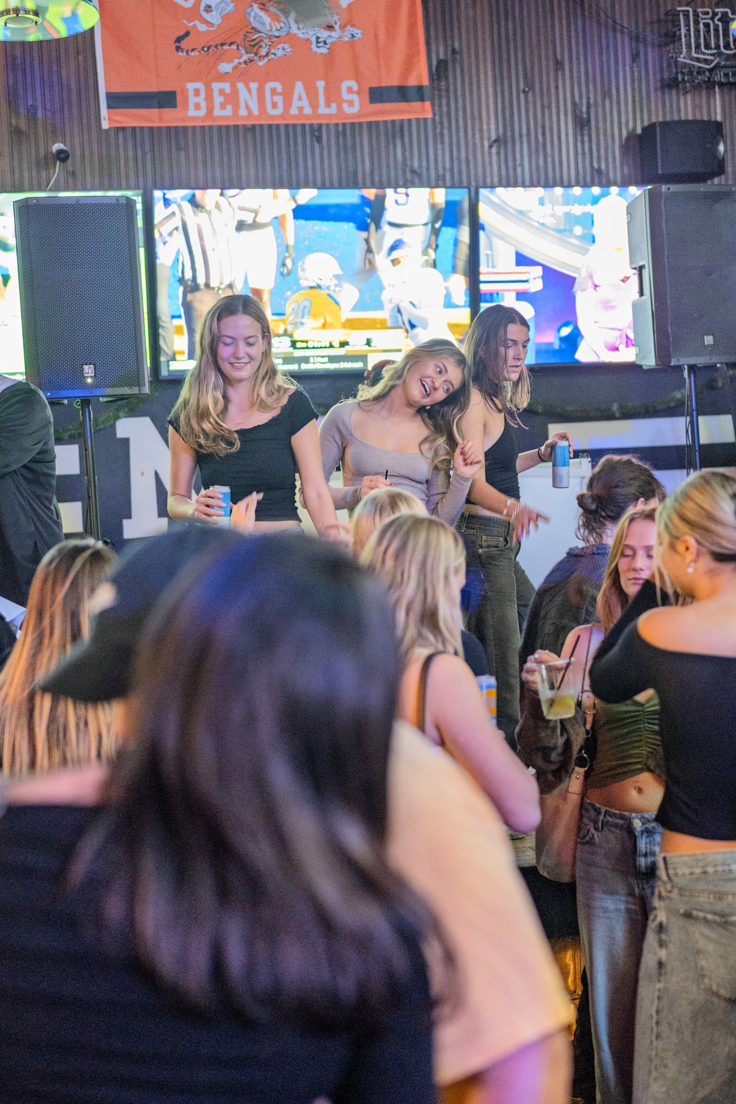 Young women dancing and socializing at a lively bar party with a Bengals banner in the background.