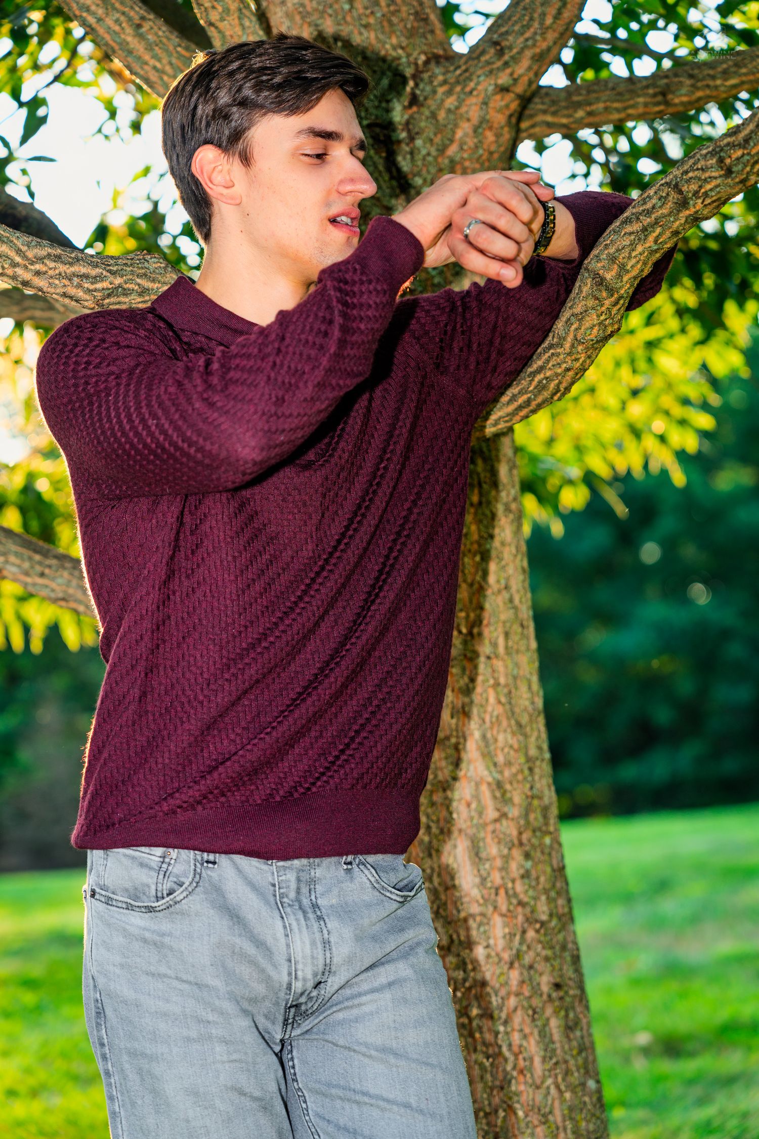Young man in burgundy sweater and jeans leaning against a tree in a sunny park, checking his watch.