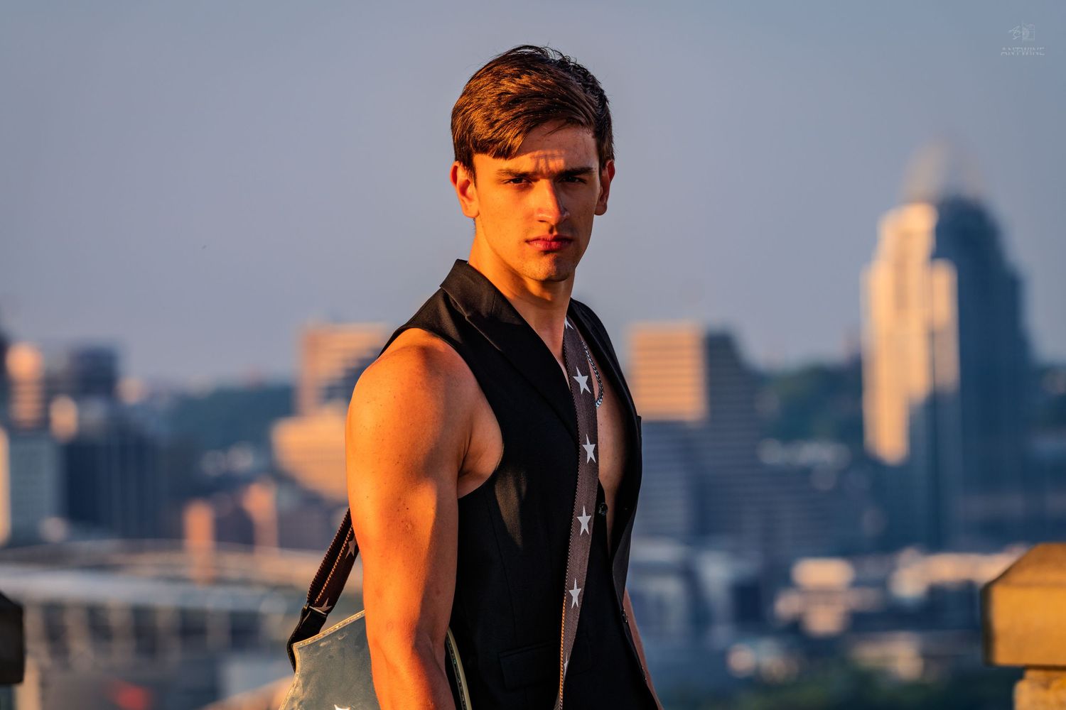 Stylish young man in sleeveless black top posing with urban skyline at sunset.