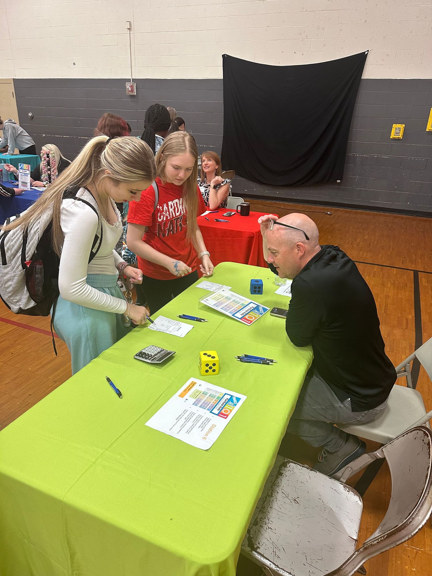Two female students engage with a bald male representative playing a dice game at a green table at a career fair.