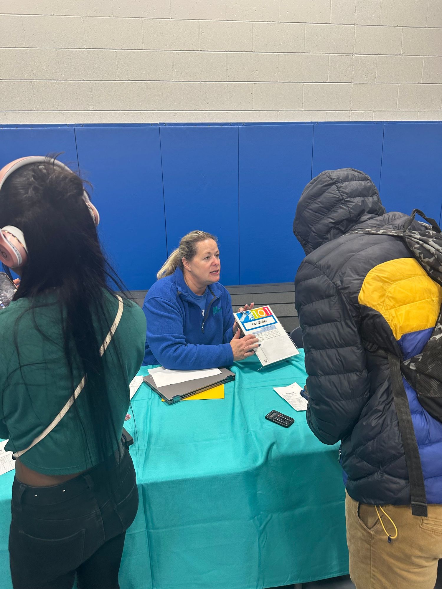 A woman in a blue jacket sits at a teal table speaking with two students at an indoor school career fair.