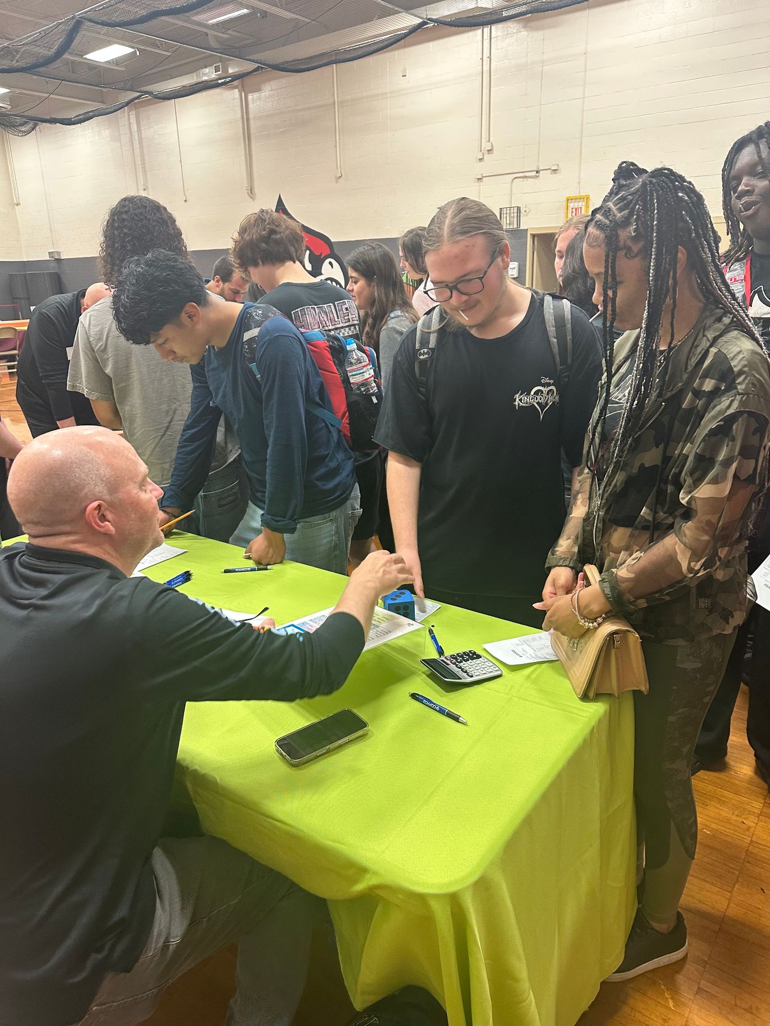 Students line up at a green table in a gym as a bald man seated signs autographs for teens at a school event.