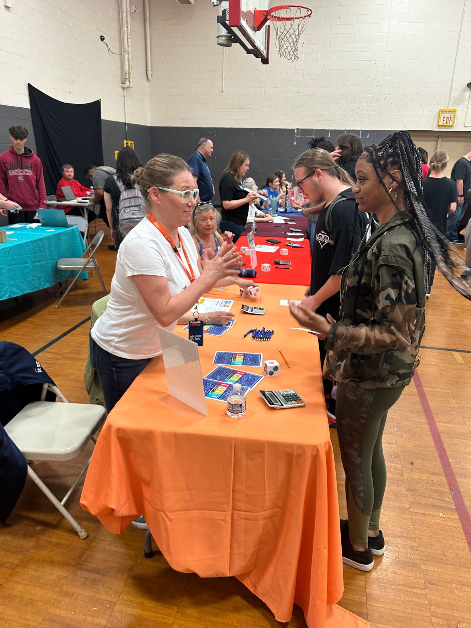 Woman in white shirt explains information at an orange-draped table to students during a school gymnasium career fair event.