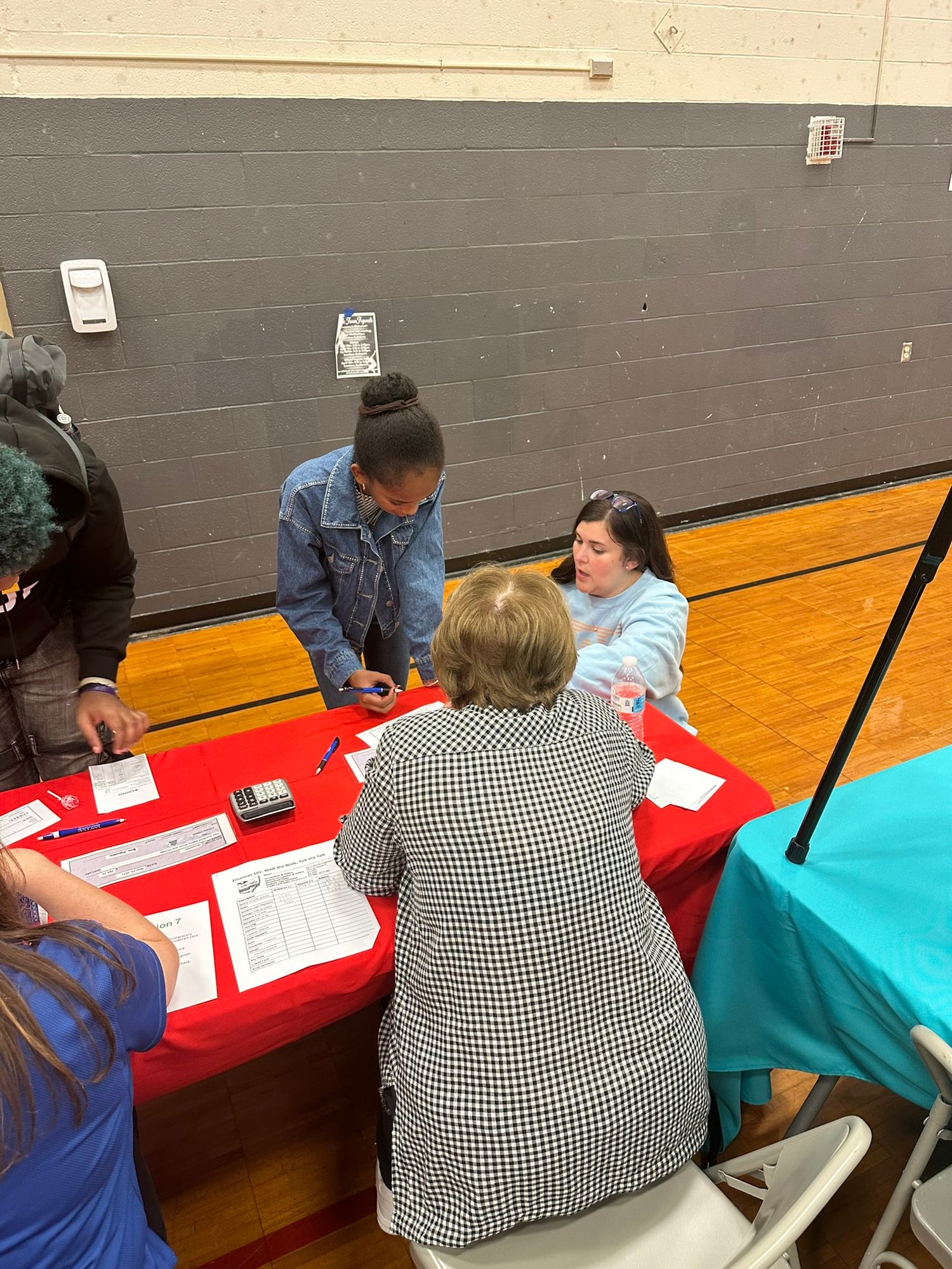 Students and adults gather at a red-covered registration table in a school gymnasium during an educational event.