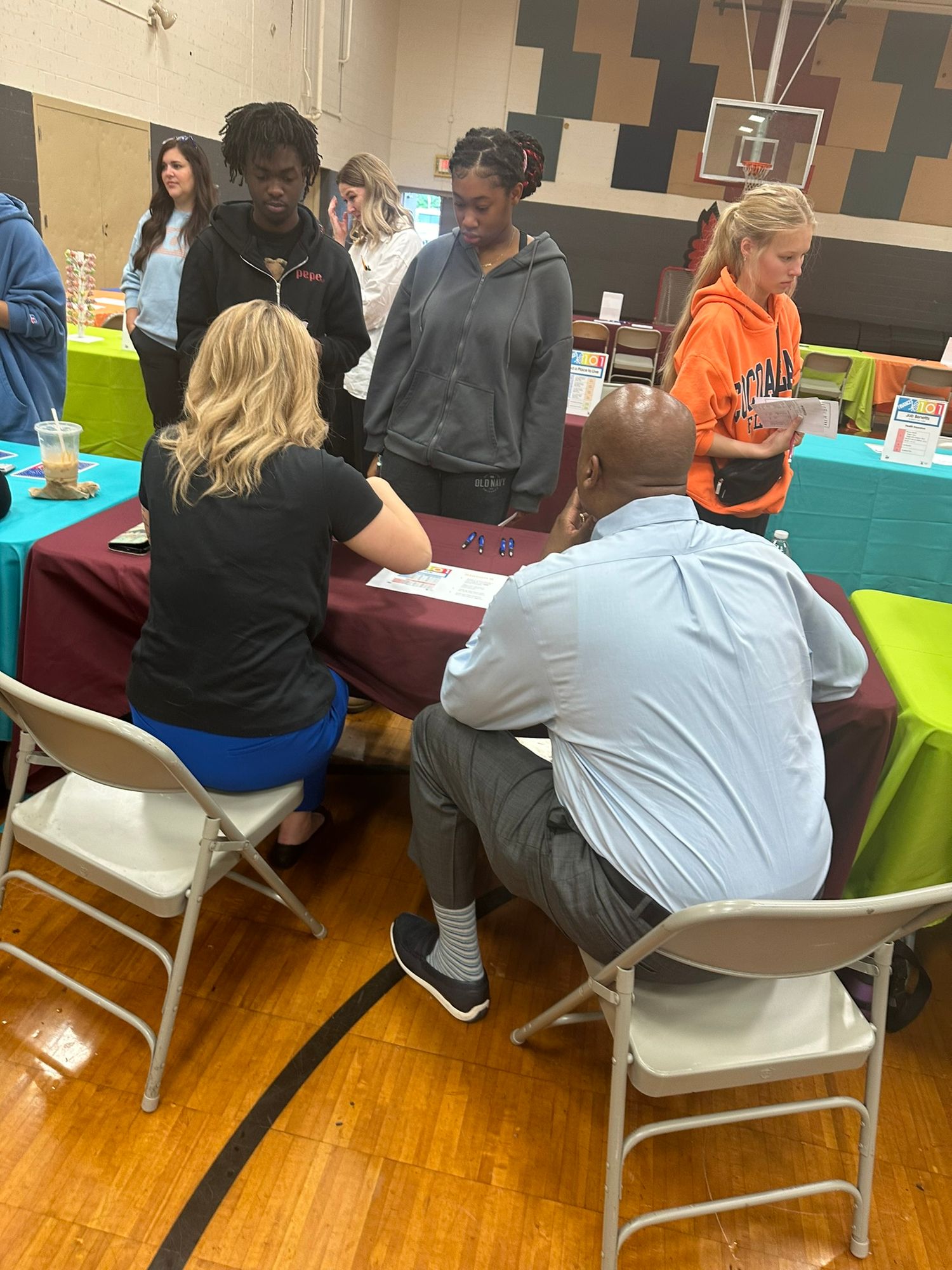 Students and adults gather around tables at an indoor school gymnasium career fair event.