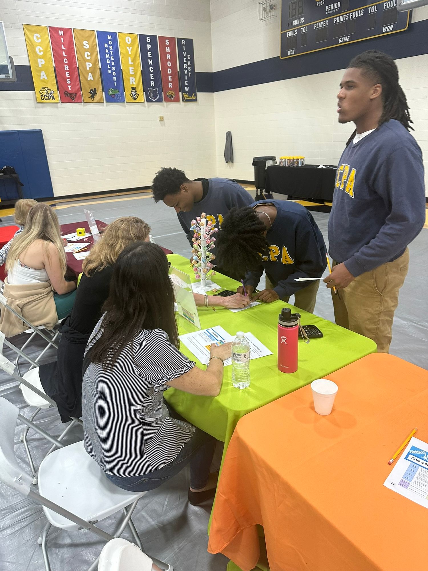 Students gather around a bright orange table in a school gymnasium during a college or career fair event.