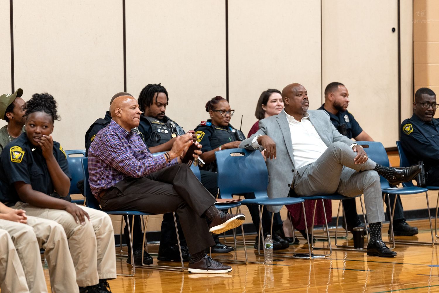 Diverse group of adults and youth seated in rows of blue chairs during a community meeting in a gymnasium.