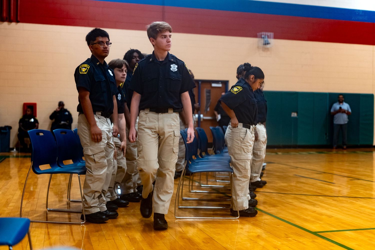 Young students in uniform shirts and khaki pants march in formation inside a school gymnasium during a JROTC drill.